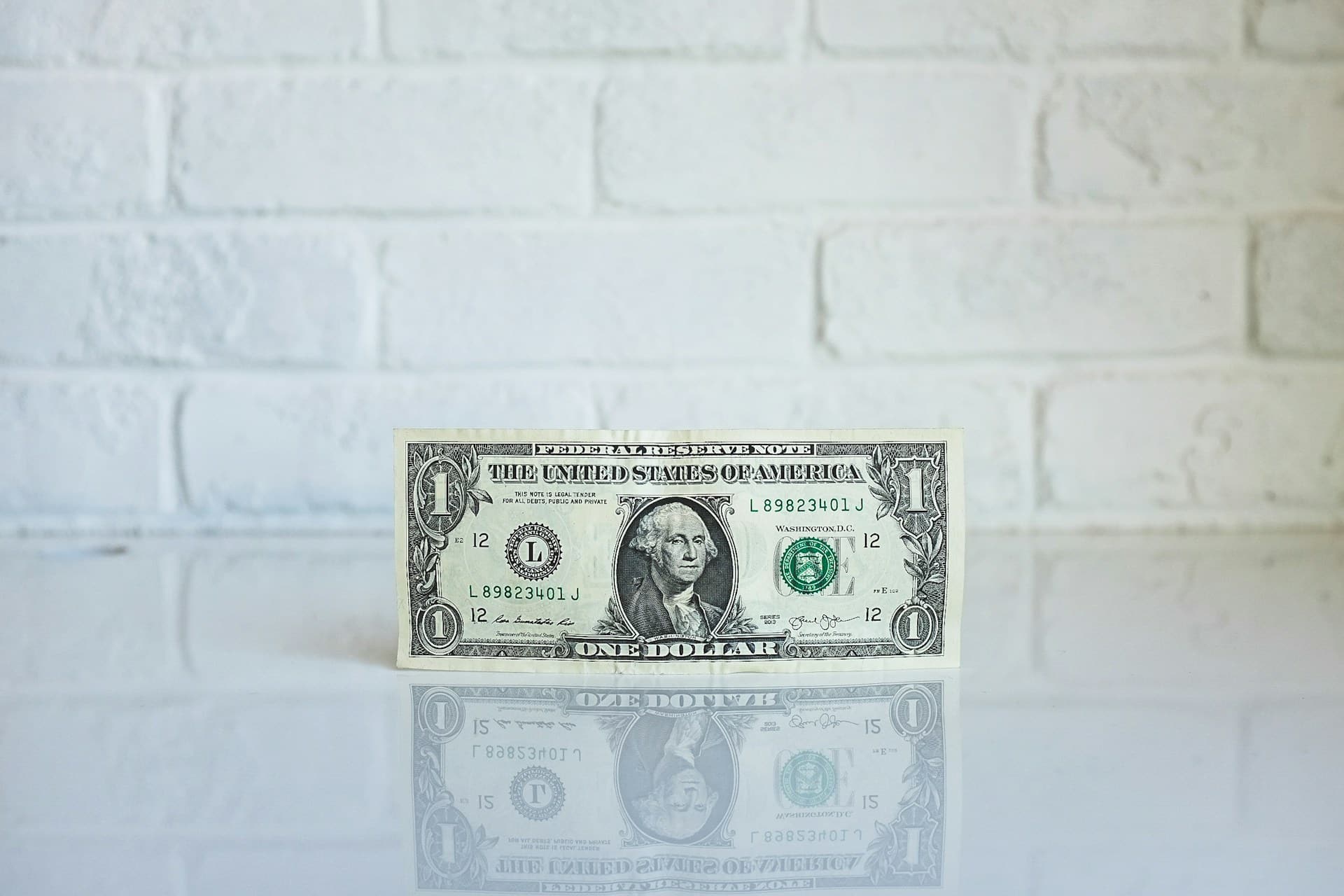 A single dollar bill with George Washington's face on it, against a white brick wall background